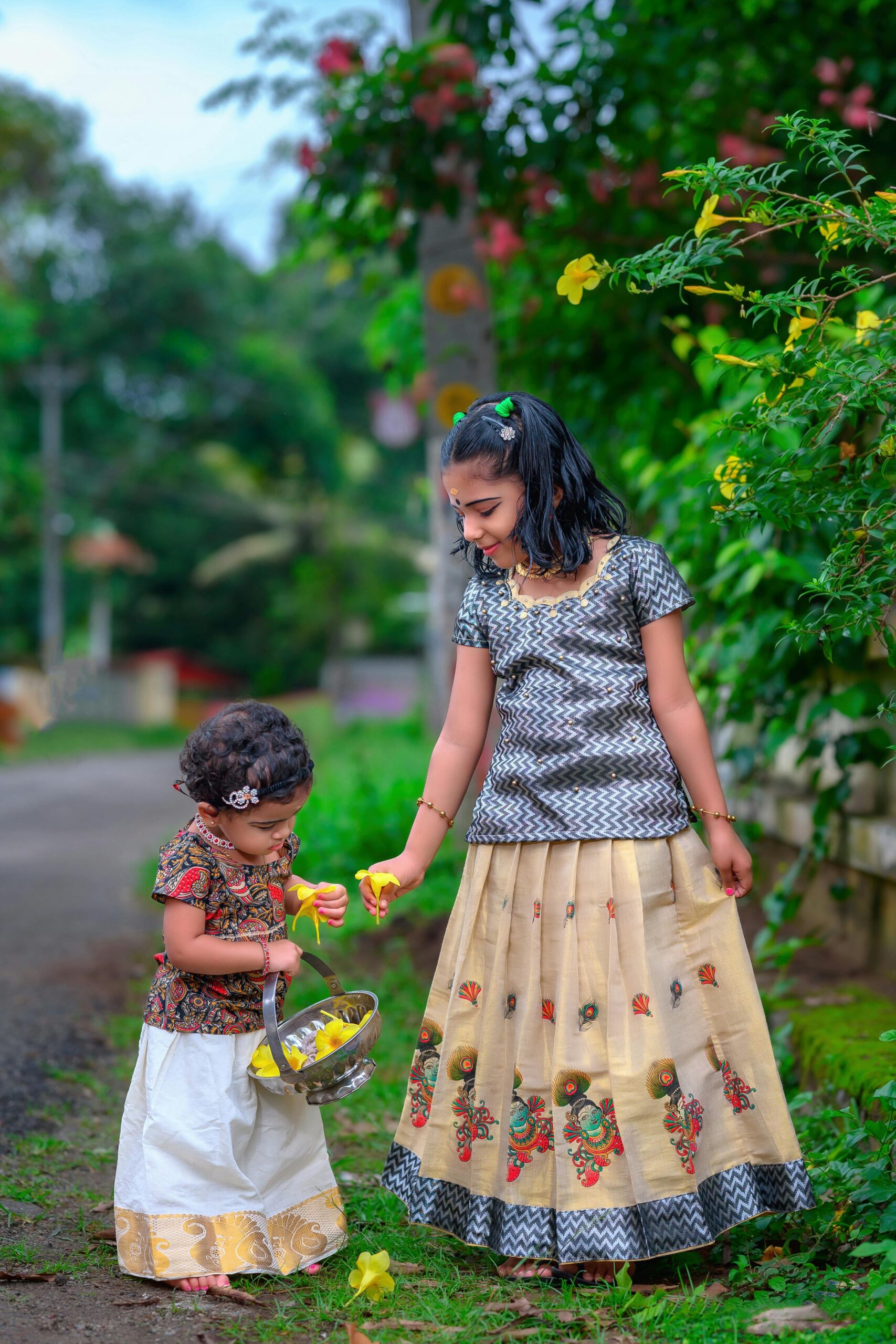 Two young girls in traditional Indian clothing gather flowers outdoors, capturing a vibrant rural setting.