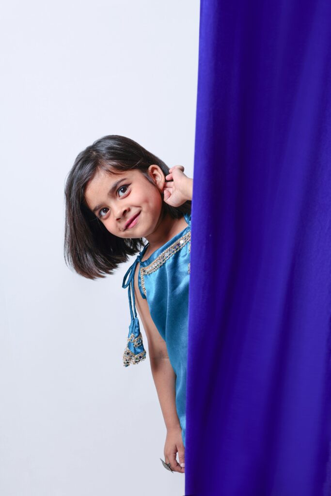 Charming young girl peeking playfully from behind a vibrant blue curtain indoors.